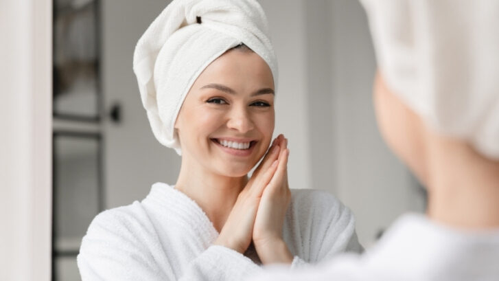 Happy woman with towel wrap smiling at mirror after dental care routine.