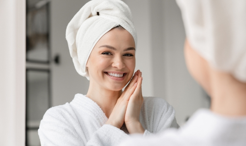 Happy woman with towel wrap smiling at mirror after dental care routine.