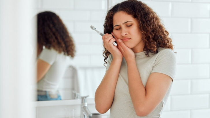 Woman applying toothpaste with brush while looking in bathroom mirror.