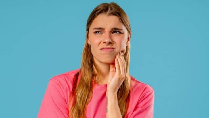 Woman in pink shirt touching her cheek in pain, depicting dental discomfort or tooth sensitivity.