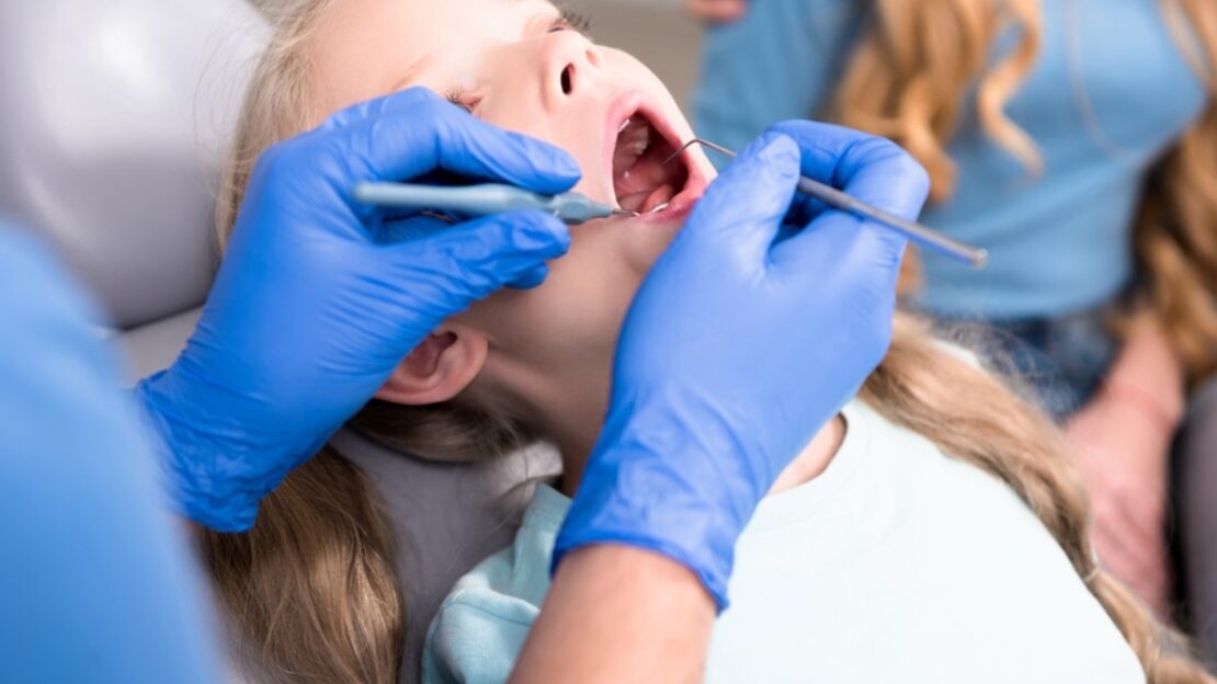 Pediatric dental exam showing a young patient receiving oral inspection with dental tools.