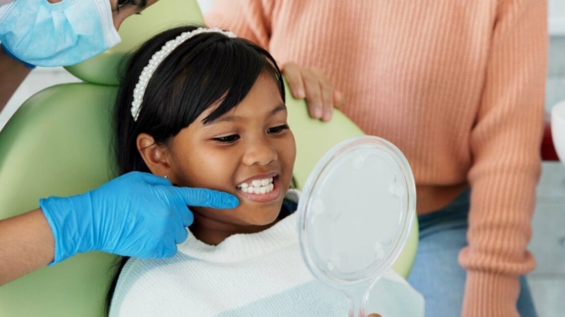 Young girl smiling at mirror during dental checkup with dentist and parent at Snellville practice.