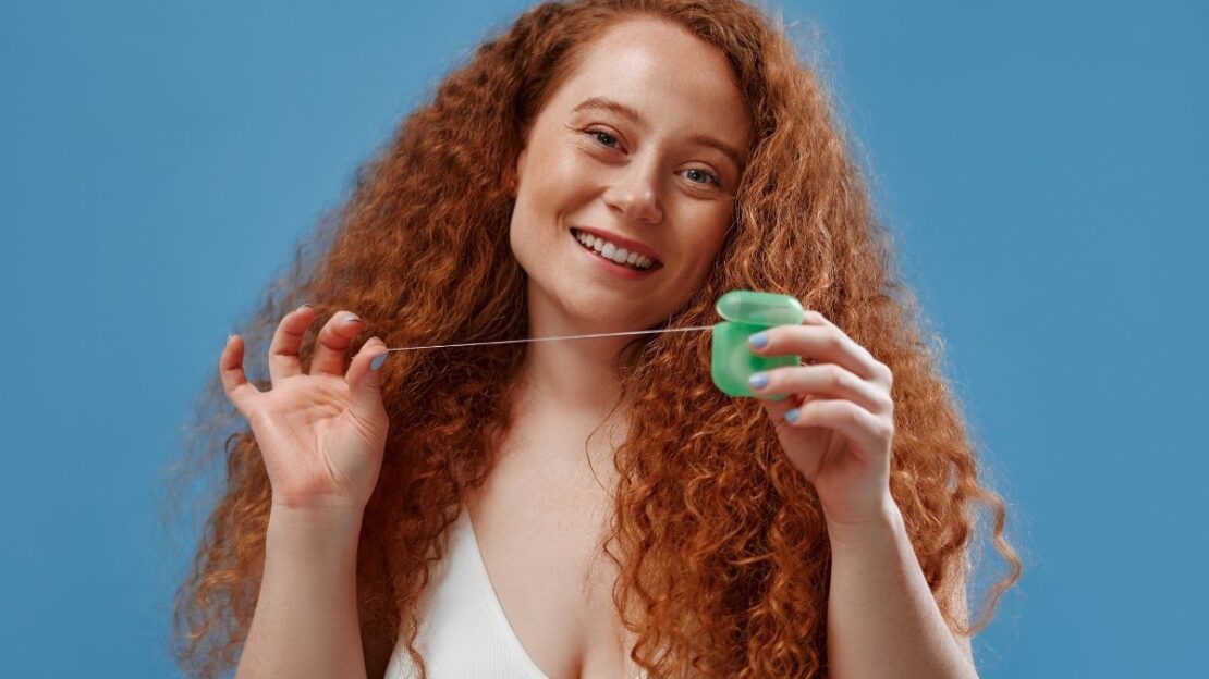 Young woman with red curly hair demonstrating proper flossing technique with green floss.