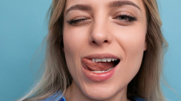 Woman with open mouth showing tongue and teeth against blue background.