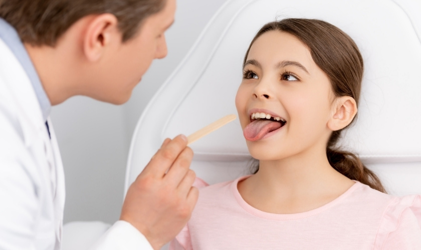 Dentist examining young patient's tongue with wooden depressor during oral examination.