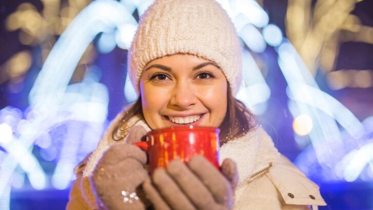 Smiling woman holding red mug wearing winter beanie and white gloves with holiday lights behind her.