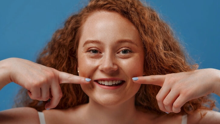 Happy young woman with red curly hair pointing to her bright smile against blue background.