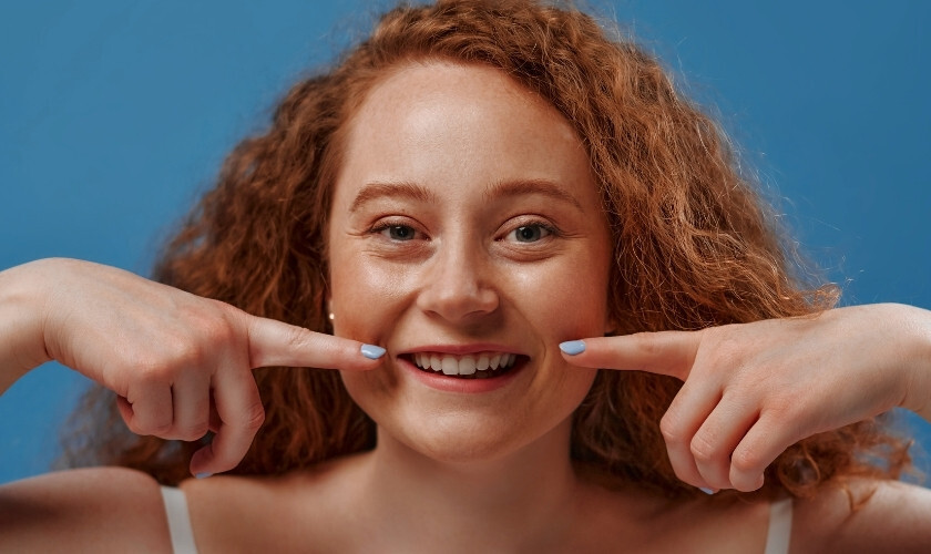 Happy young woman with red curly hair pointing to her bright smile against blue background.