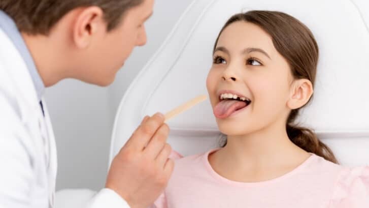 Dentist examining child patient's tongue with wooden depressor during oral examination.