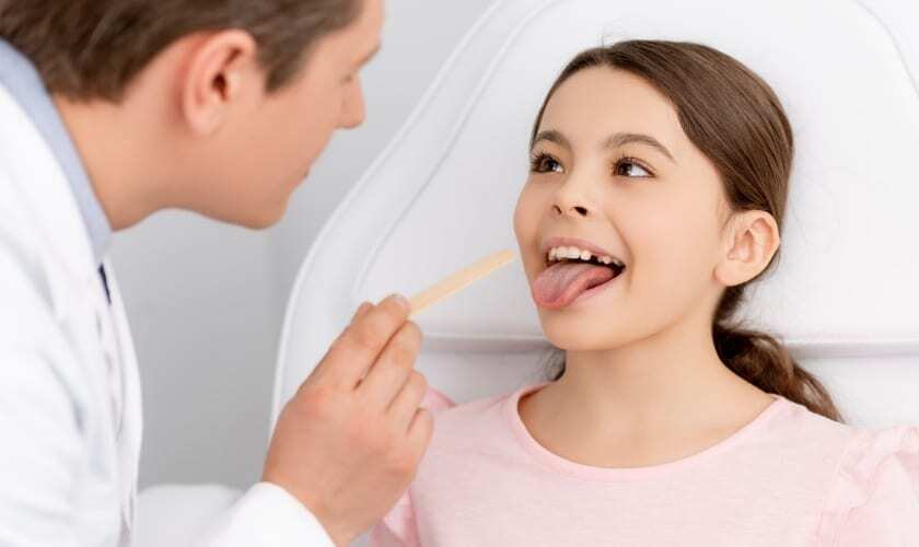Dentist examining child patient's tongue with wooden depressor during oral examination.