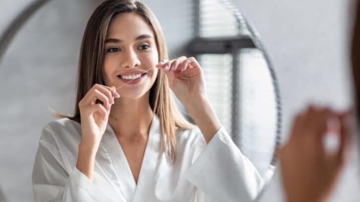 Woman flossing teeth with string floss in bathroom mirror.