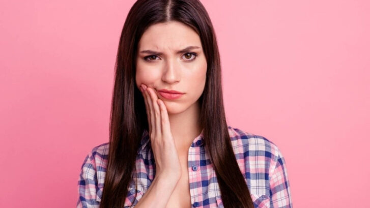 Woman with tooth pain touching her cheek against pink background.