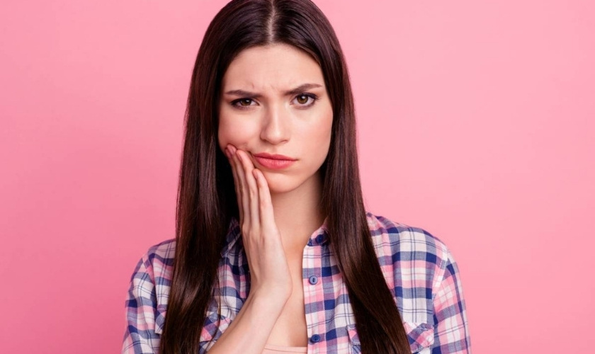 Woman with tooth pain touching her cheek against pink background.