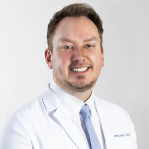 Professional male dentist in white coat and tie smiling at camera, headshot portrait.