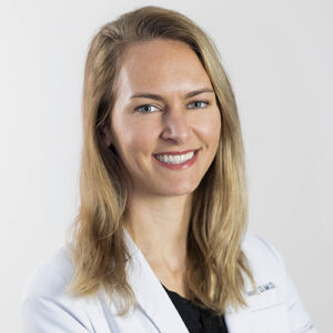 Professional female dentist in white coat smiling at camera against neutral background.