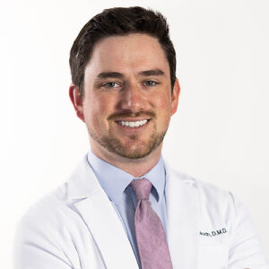Male dentist in white coat and purple tie smiling at camera in professional headshot.